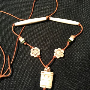 May include: A brown leather cord necklace with white bone beads. The necklace features two long bone beads, two flower-shaped beads, and a rectangular bone bead with a carved design.