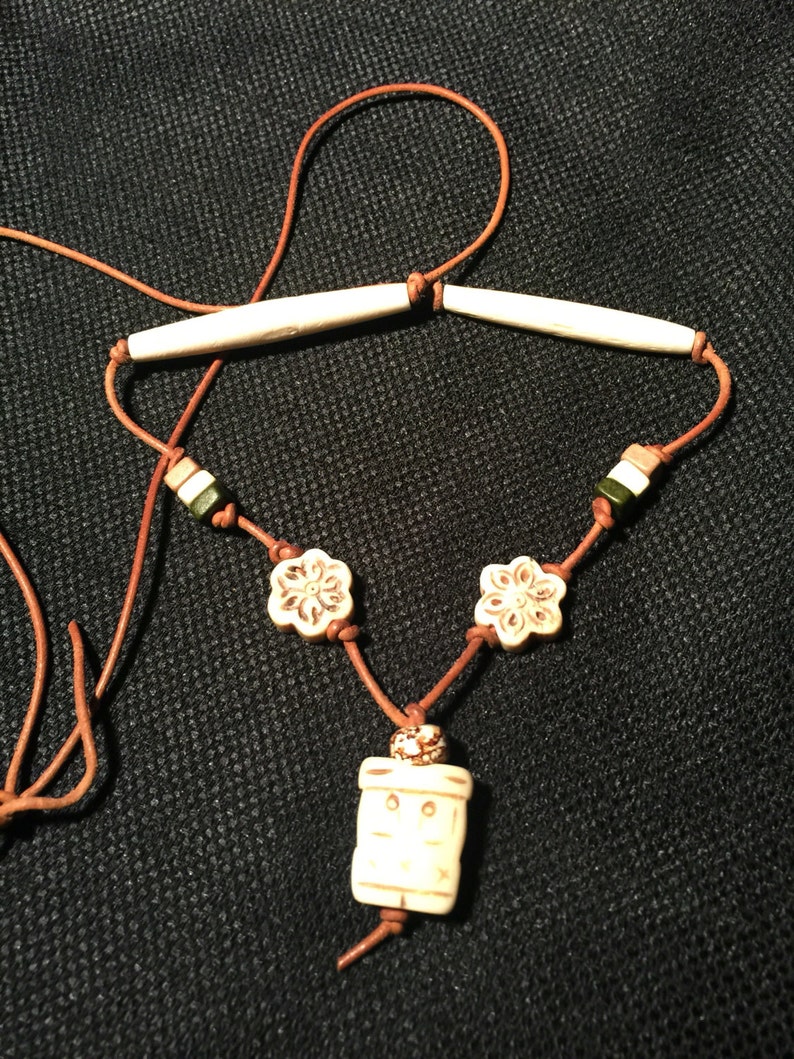 May include: A brown leather cord necklace with white bone beads. The necklace features two long bone beads, two flower-shaped beads, and a rectangular bone bead with a carved design.