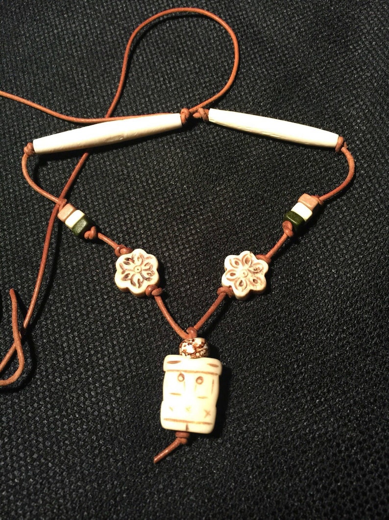 May include: A brown leather cord necklace with a bone pendant and bone beads. The pendant is rectangular with a carved design. The beads are round and have a floral design.