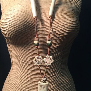 May include: A brown leather necklace with white bone beads, green and brown square beads, and two white bone flower charms. The necklace has a white bone pendant with a carved design.