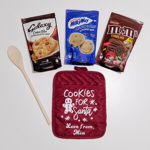 May include: A red oven mitt with the text "Cookies for Santa" and a gingerbread man graphic. Three bags of cookie mix are included: Galaxy Chocolate Chip, Milky Way Magic Stars, and M&M's Chocolate.