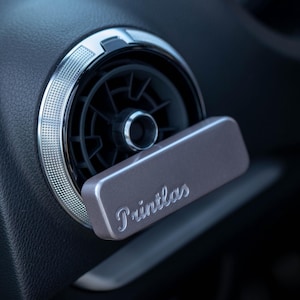 Könnte beinhalten: Nahaufnahme einer Auto-Luftdüse mit silberner Zierleiste. Ein rechteckiger, taupefarbener Lufterfrischer mit dem Wort "Printlas" in Schreibschrift ist in die Düse eingesetzt. Die Düse befindet sich vor einem dunkelgrauen Armaturenbrett.