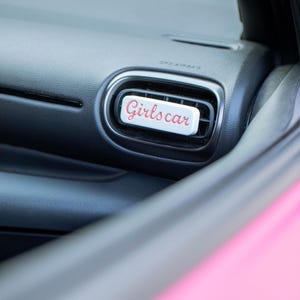 Könnte beinhalten: Ein weißer Lufterfrischer für das Auto mit dem Text "Girls car" in rosa Schrift. Der Lufterfrischer ist rechteckig und sitzt in einer schwarzen Lüftungsöffnung des Autos. Das Armaturenbrett des Autos ist schwarz und das Äußere des Autos ist rosa.