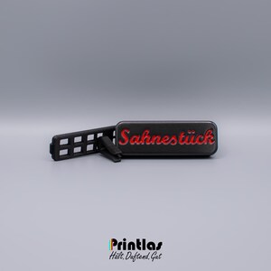 Pode incluir: Um ambientador retangular preto com a palavra "Sahnestück" em escrita vermelha. O ambientador tem um clip preto e um suporte preto perfurado. A parte inferior da imagem tem o texto "Printlas" e "Hält. Duftend. Gut."
