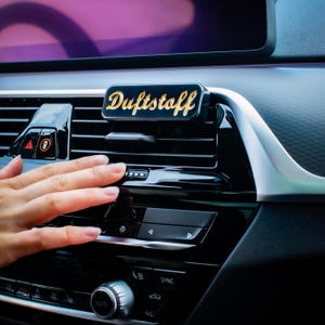 Op de afbeelding: Een zwarte auto luchtverfrisser met gouden letters "Duftstoff" is bevestigd aan een ventilatierooster in de auto. Het interieur van de auto is zwart met zilveren accenten. Een hand reikt naar het ventilatierooster.