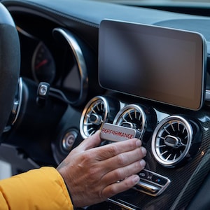 May include: A silver rectangular car vent accessory with the word "PERFORMANCE" in red lettering. The accessory is being held in front of a car's dashboard, near the air vents and a digital display.