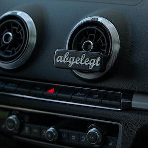 Puede incluir: Accesorio negro para la ventilación del coche con la palabra "abgelegt" en escritura plateada. El accesorio rectangular está montado en el salpicadero de un coche cerca de la ventilación.