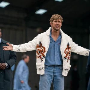 May include: A cream-colored knit cardigan featuring a brown and orange fox graphic on the front panels, worn over a blue button-down shirt and light blue denim jeans. The cardigan has a shawl collar, ribbed cuffs, and two front pockets.