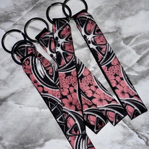May include: Four black and pink keychains with silver key rings. The keychains feature a repeating pattern of pink flowers and black wheels.