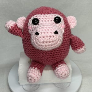 May include: A handmade crochet monkey doll in shades of pink. The monkey has a round body, large black eyes, and a light pink face, ears, hands, and feet. The doll is sitting on a white surface.