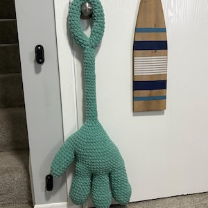 May include: A large, teal-colored, crocheted monster hand toy with a long stem and loop for hanging. The hand has five fingers. A wooden oar with blue and white stripes hangs on the door behind the toy.