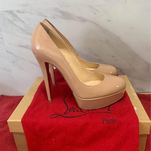 May include: A pair of nude patent leather platform pumps with stiletto heels. The shoes are displayed on a box with a red dust bag. The box has the word "Paris" printed on it.
