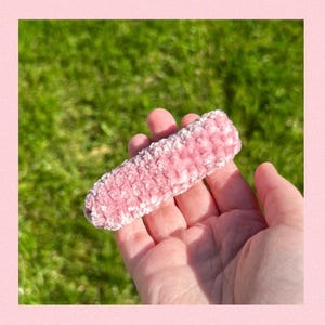 May include: A pink fuzzy hair clip. The clip is held in a hand against a green grassy background.