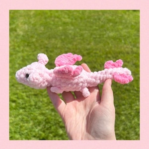 Puede incluir: Un dragón de crochet rosa con una textura esponjosa. El dragón tiene alas y una cola larga.