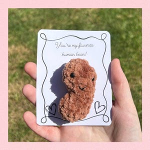 Puede incluir: Un juguete de peluche en forma de judía marrón con una cara sonriente, sujeto a una tarjeta blanca. La tarjeta tiene el texto "You're my favorite human bean!" y dos garabatos de corazón. El fondo es verde borroso.