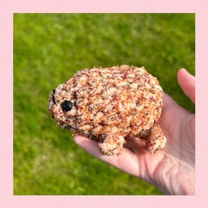 May include: A small, crocheted animal toy in shades of brown and orange. The toy has a round body and small legs. It is being held in a person's hand.
