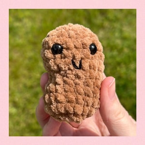 Puede incluir: Un juguete de peluche de cacahuete de punto marrón con una cara sonriente y ojos negros.