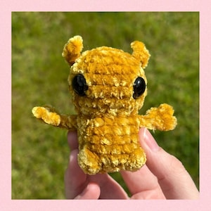 May include: A small, crocheted plush toy in a mustard yellow color. The toy has large black eyes and a textured, fuzzy surface.