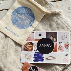 May include: A white linen bag with a small, white drawstring bag inside. The drawstring bag has a blue circle with the text "Lumafina" and the text "Made in Portland, OR". A card with a collage of images and the text "Lumafina" is on top of the bag.