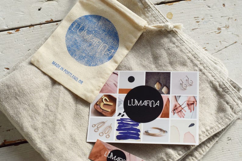 May include: A white linen bag with a small, white drawstring bag inside. The drawstring bag has a blue circle with the text "Lumafina" and the text "Made in Portland, OR". A card with a collage of images and the text "Lumafina" is on top of the bag.