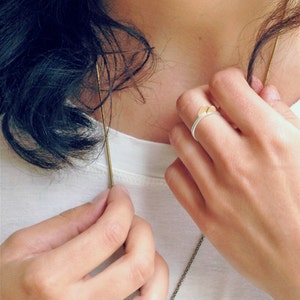 May include: A woman wearing a white shirt with a gold chain necklace and two rings on her fingers. One ring is a gold band with a clear stone, and the other is a silver band with a gold accent.