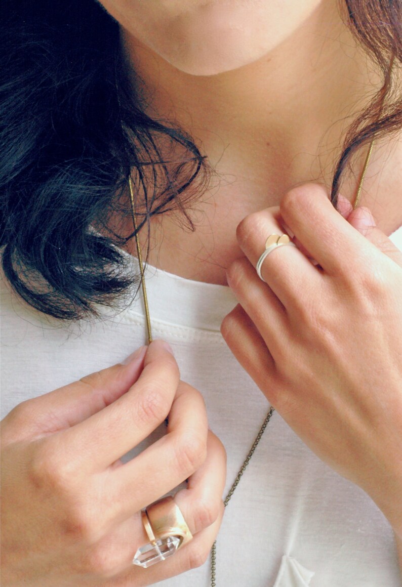 May include: A woman wearing a white shirt with a gold chain necklace and two rings on her fingers. One ring is a gold band with a clear stone, and the other is a silver band with a gold accent.