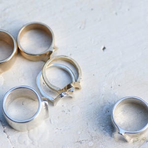May include: A collection of six silver and gold rings with unique designs. The rings are arranged on a white surface.