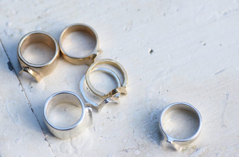 May include: A collection of six silver and gold rings with unique designs. The rings are arranged on a white surface.