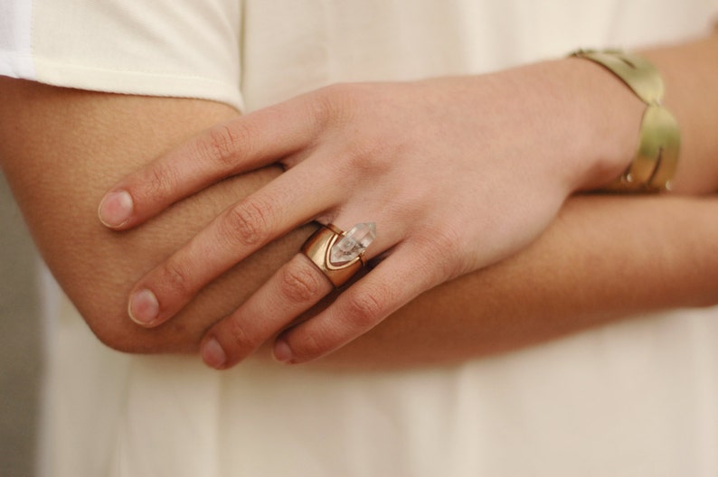 May include: A gold ring with a clear quartz crystal point set in a gold band. The ring is worn on the index finger of a person's hand.