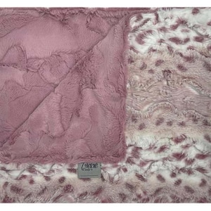 May include: A soft, pink baby blanket with a textured surface. One side of the blanket is a solid pink, while the other has a white and pink animal print. A small label with the brand name is visible.