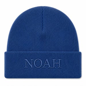 Puede incluir: Un gorro de punto azul real con un dobladillo plegado y la palabra NOAH bordada en hilo azul a juego en la parte delantera. El gorro presenta una textura acanalada vertical en toda la corona y un acabado de punto liso en el dobladillo.