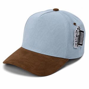 Può includere: Un cappellino da baseball azzurro con visiera effetto camoscio marrone e un bottone marrone sulla corona. Un'etichetta rettangolare bianca attaccata al lato riporta la scritta BASIC CAP in nero, insieme a un codice a barre e altro testo.