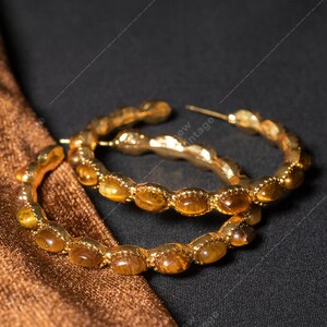 May include: Gold-toned hoop earrings with amber-colored stones. The earrings are set against a dark background with a brown fabric. The earrings have a vintage aesthetic.