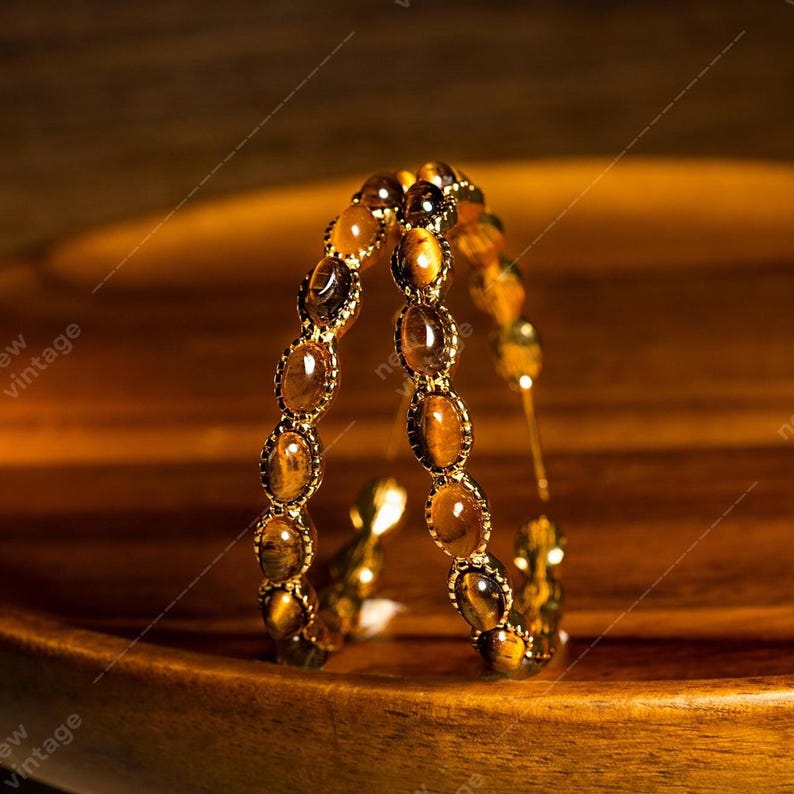 May include: A pair of gold-toned bracelets with oval-shaped tiger's eye stones. The stones are set in a gold metal frame. The bracelets are displayed on a wooden surface, creating a warm, natural aesthetic. The bracelets are a jewelry item.