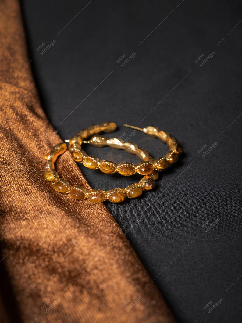 May include: Gold-toned hoop earrings with amber-colored stones. The earrings are set against a dark background with a brown fabric. The earrings have a vintage aesthetic.