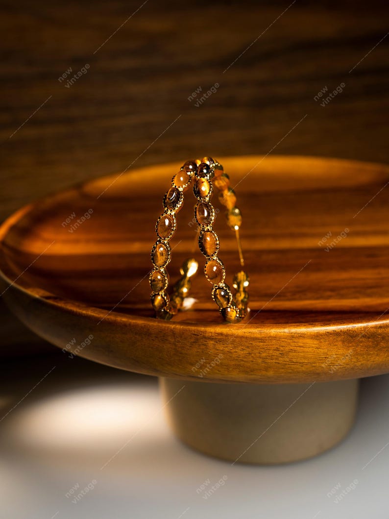 May include: A gold-toned bracelet with oval-shaped tiger's eye stones. The bracelet is displayed on a wooden pedestal with a round top. The background is a warm, neutral tone.