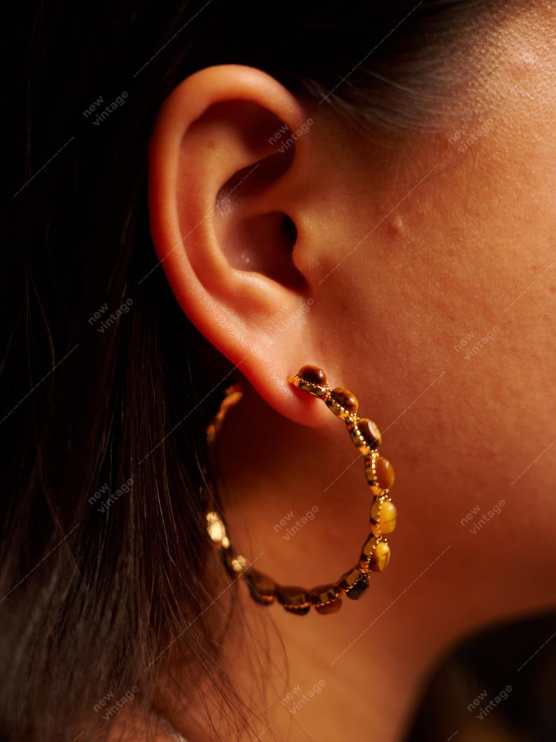 May include: Gold-toned hoop earrings with a series of small, amber-colored stones. The earrings are worn on an ear, with the skin tone visible. The earrings have a vintage aesthetic.