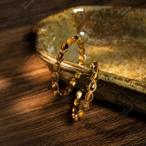 May include: A gold-toned bracelet with oval-shaped tiger's eye stones. The bracelet is displayed on a rustic, ceramic dish with a brown wooden surface in the background. The bracelet is a piece of jewelry.