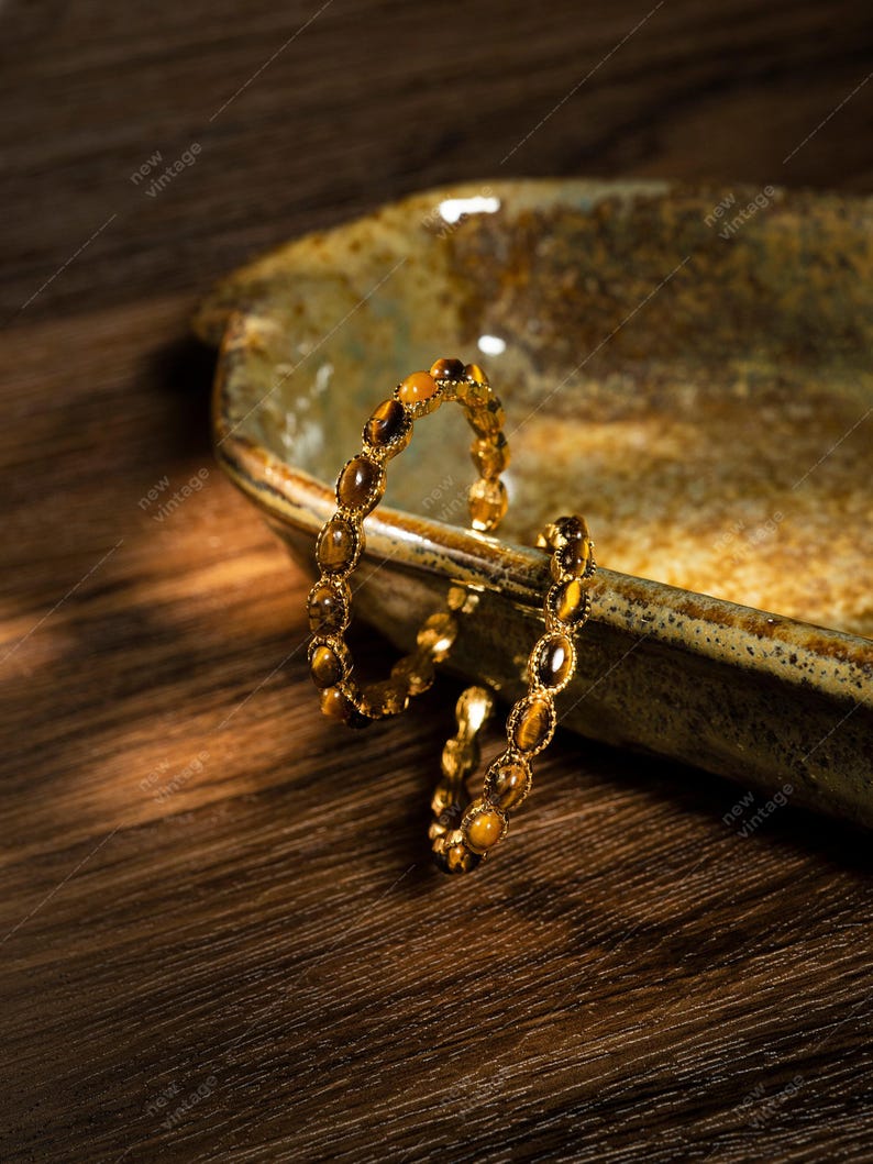May include: A gold-toned bracelet with oval-shaped tiger's eye stones. The bracelet is displayed on a rustic, ceramic dish with a brown wooden surface in the background. The bracelet is a piece of jewelry.