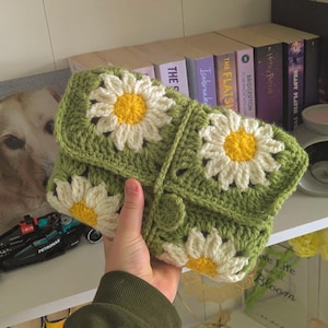 rocheted book cover featuring daisy granny squares with white petals and yellow centres set against a green background. Handmade, floral, and cozy book protection