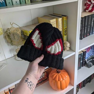 May include: A black crocheted beanie featuring two prominent, stylised eye designs. Each eye is crafted with a white centre and a bold red outline, creating a distinct graphic appearance. The hat has a thick, ribbed cuff at the base for a secure fit.