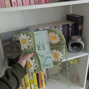 rocheted book cover featuring daisy granny squares with white petals and yellow centres set against a green background. Handmade, floral, and cozy book protection