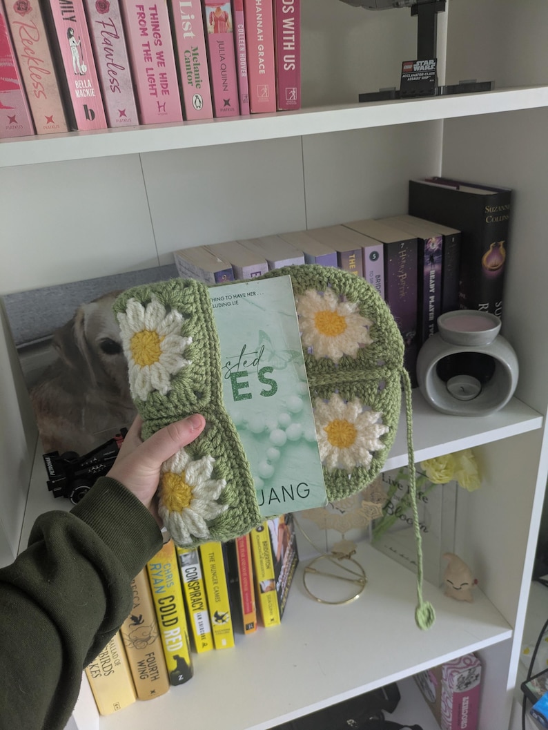 rocheted book cover featuring daisy granny squares with white petals and yellow centres set against a green background. Handmade, floral, and cozy book protection