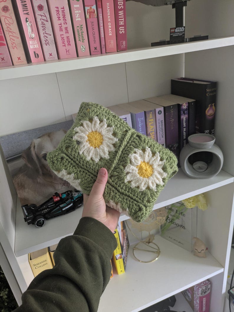 rocheted book cover featuring daisy granny squares with white petals and yellow centres set against a green background. Handmade, floral, and cozy book protection