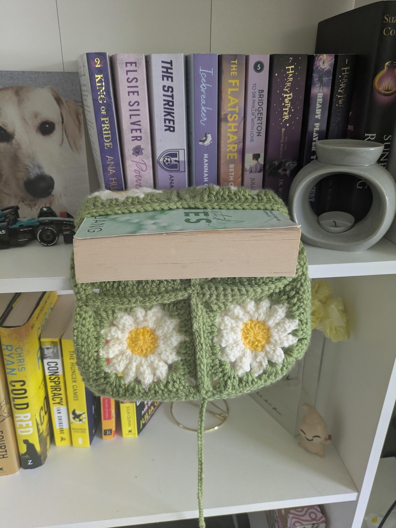 rocheted book cover featuring daisy granny squares with white petals and yellow centres set against a green background. Handmade, floral, and cozy book protection