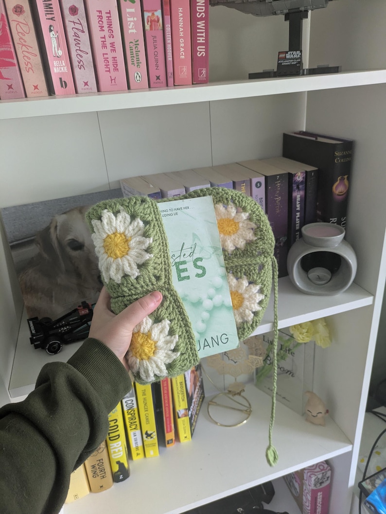 rocheted book cover featuring daisy granny squares with white petals and yellow centres set against a green background. Handmade, floral, and cozy book protection