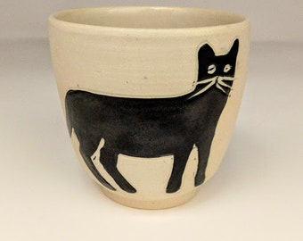 Puede incluir: Una taza de cerámica blanca con una silueta negra de un gato en el lateral. El gato está mirando hacia la izquierda y tiene un diseño simple.