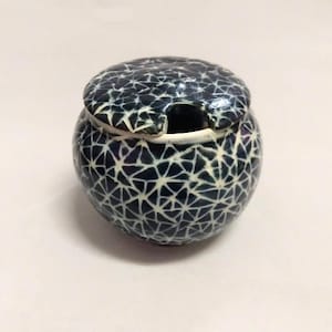 Può includere: Un piccolo barattolo rotondo in ceramica con un coperchio. Il barattolo e il coperchio sono decorati con un motivo geometrico bianco su sfondo blu scuro.