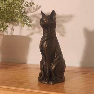 May include: Bronze cat figurine in a seated pose, featuring a streamlined design. The cat statue is placed on a wooden shelf, with a plant in a white pot in the background. The shelf is supported by a gold-coloured bracket.