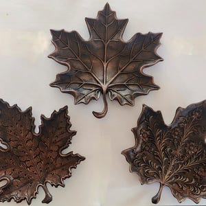 May include: Three bronze-finished maple leaf-shaped trays. The trays feature detailed veining and floral patterns. The largest tray is centered, with two smaller trays below. These trays are ideal for autumn decor.